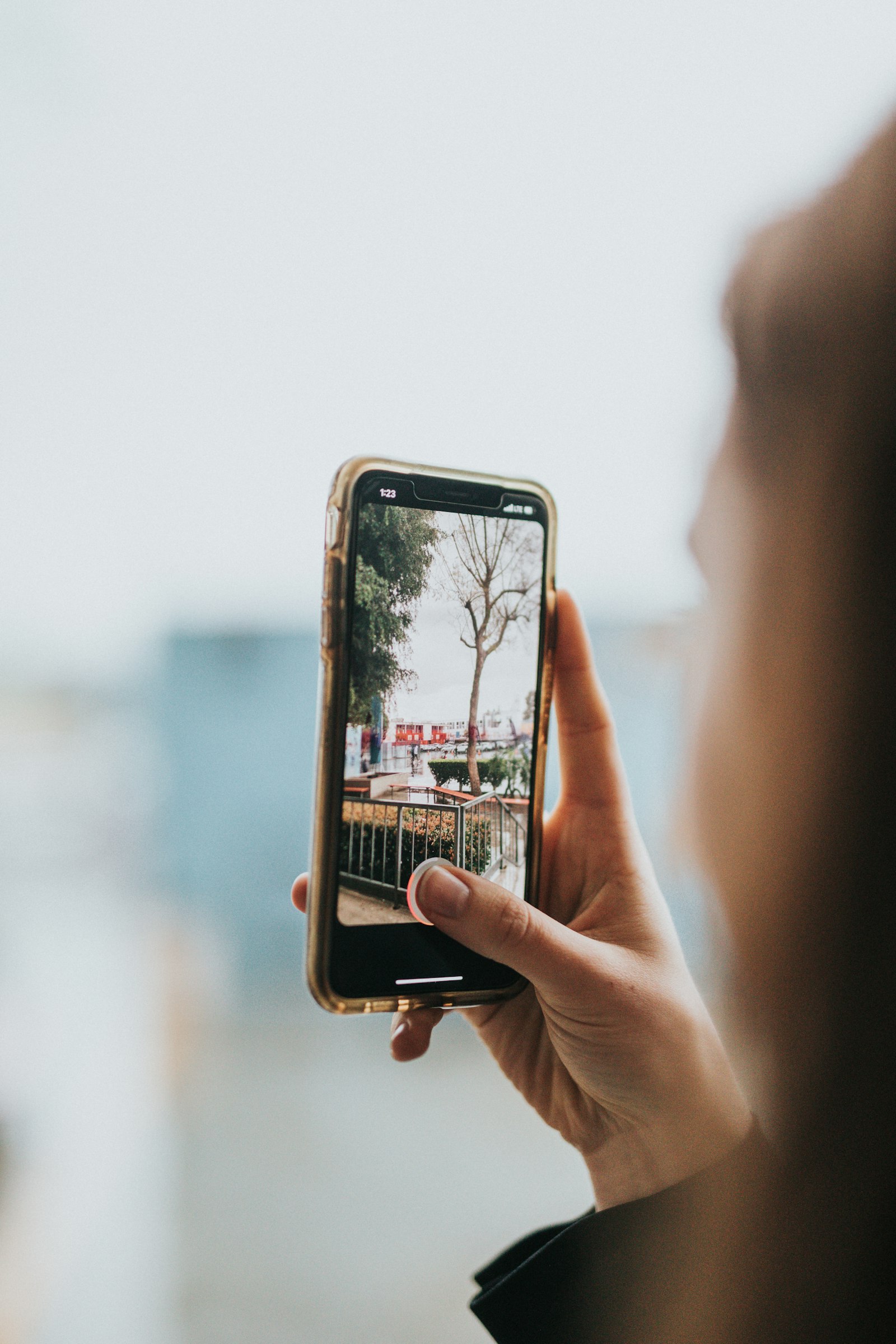 A person holding up a smartphone, taking a photo of a tree-lined waterfront